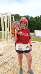Emily holding a nail gun while posing near an unfinished house frame